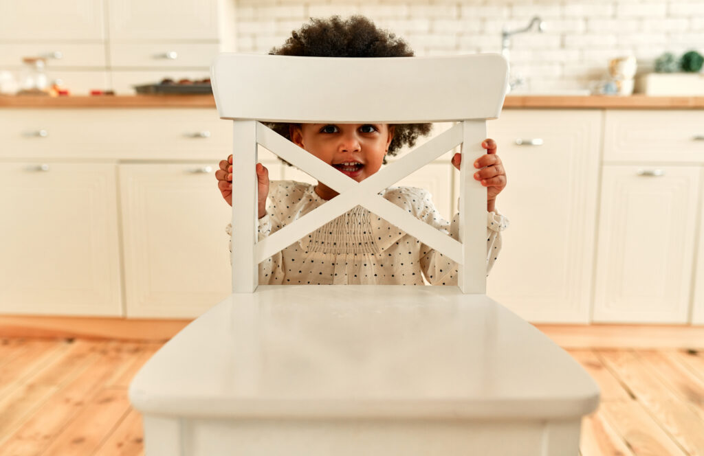 A cute African American child girl with curly fluffy hair stands behind an armchair and looks out cheerfully smiling in the kitchen at home.